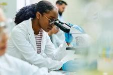 A Black woman in a lab coat peers through the lens of a microscope in a lab.