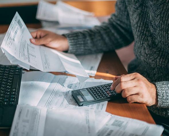 A man sits at a table in front of a computer with a calculator, going through various bills.